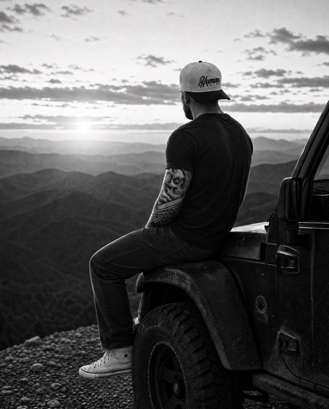 Sitting on the fender of a parked 4x4 at a mountain overlook at sunset, HUMAN BEING cap